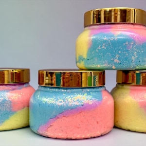 May include: Four clear plastic jars with shiny gold lids, each filled with a swirled mixture of pastel pink, blue, and yellow cream. The contents have a textured, glittery appearance, and the jars are arranged in a stacked formation.