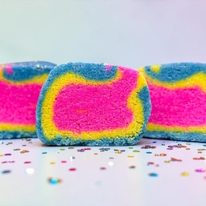 Puede incluir: Cinco bombas de baño coloridas y en capas con un diseño vibrante. Cada bomba de baño presenta un centro rosa brillante, rodeado por una capa amarilla y una capa exterior azul. Las bombas de baño están sobre una superficie blanca con confeti colorido.
