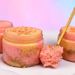 May include: Three jars of pink and tan body scrub with a golden spoon. The scrub has a layered appearance and is topped with small, golden sprinkles. The jars are displayed on a white surface against a pastel background.