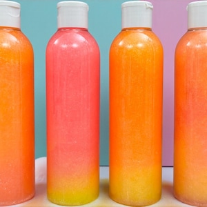 Op de afbeelding: Vier transparante plastic flessen gevuld met een kleurrijke vloeistof, met een verloop van oranje, roze en geel. Elke fles heeft een witte dop. De flessen staan in een rij, mogelijk verzorgingsproducten.