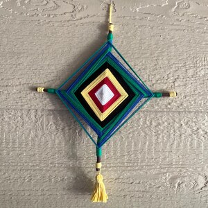 Peut inclure: Un objet décoratif en forme de losange composé de fils de laine enroulés autour de deux bâtons en bois croisés. Le motif présente des couches concentriques de fils blanc, rouge, jaune, noir, vert et bleu, avec un pompon jaune en bas.