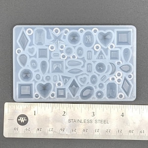 May include: A clear, rectangular silicone mold filled with various gemstone shapes, including diamonds, hearts, and ovals. A stainless steel ruler with inch markings is placed next to the mold for scale. The mold is designed for crafting jewelry or decorative elements.