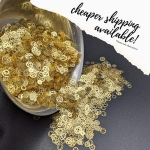 May include: A collection of small, gold-colored watch gears spilling from a metal container onto a dark surface. The gears vary in size and design, creating a detailed and intricate pattern. Text in the image reads "cheaper shipping available!"
