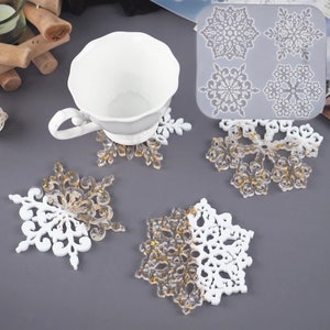 May include: White and clear snowflake-shaped coasters with gold flecks, and a white teacup. A silicone mold with snowflake designs is also visible. These decorative coasters are perfect for winter or holiday decor.