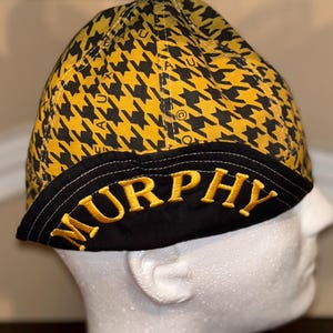 May include: A black and yellow houndstooth patterned cap with the name "MURPHY" embroidered in gold. The cap is displayed on a white mannequin head.