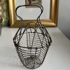 Op de afbeelding: Vintage draadmand met een handvat. De mand is gemaakt van dunne, zilverkleurige metalen draden, geweven tot een afgeronde vorm. Het handvat loopt over de bovenkant. De mand wordt waarschijnlijk gebruikt voor het dragen van kleine voorwerpen of als decoratief stuk.
