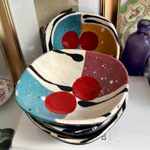 Puede incluir: Conjunto de cuatro cuencos de cerámica con un diseño geométrico. Los cuencos están pintados en tonos de azul, rojo, amarillo, negro y blanco. Cada cuenco tiene dos círculos rojos en el centro.