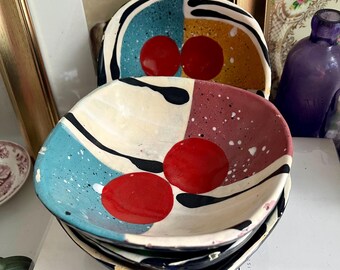 Pode incluir: Pilha de quatro tigelas de cerâmica com um design geométrico. As tigelas são pintadas em tons de azul, vermelho, amarelo, preto e branco. Cada tigela tem dois círculos vermelhos no centro.