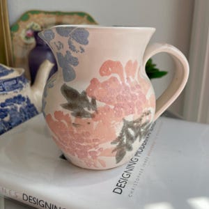 Puede incluir: Una jarra de cerámica blanca con un diseño floral en tonos rosa, azul y gris. La jarra tiene una boca ancha y un asa.