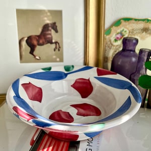 Puede incluir: Un cuenco de cerámica blanca con un diseño abstracto en azul, verde y rojo. El cuenco está ligeramente inclinado y tiene una forma redondeada.