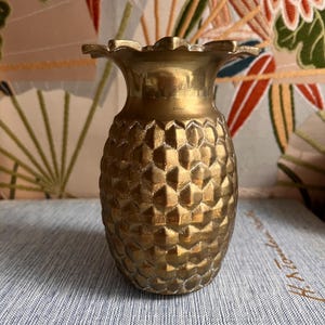 May include: A metallic gold-toned vase shaped like a pineapple, featuring a textured, diamond-patterned exterior and a flared, scalloped rim. The object has a polished finish with subtle variations in colour, reflecting light across its surface.
