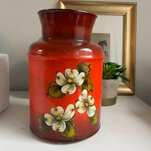 May include: A red and orange ceramic pitcher with a glossy finish. The pitcher features hand-painted white flowers with yellow centers and green leaves. The top of the pitcher is a darker shade of red.