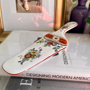 Puede incluir: Un servidor de pastel de cerámica blanca con un diseño floral rojo, amarillo y verde. El servidor tiene un borde rojo y un mango largo y delgado.