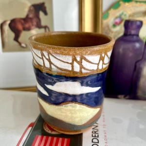 Peut inclure: Une tasse en céramique faite à la main avec un bord brun et un glaçage bleu et blanc. La tasse a un motif de paysage avec des nuages blancs et des collines brunes et blanches.