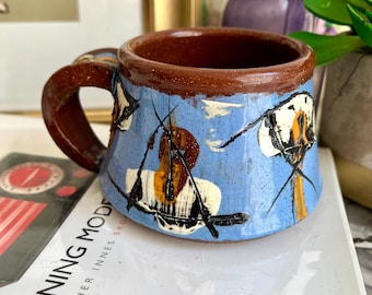 Puede incluir: Una taza de cerámica azul y marrón con asa. La taza tiene un diseño pintado de formas y líneas abstractas.