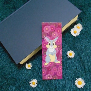 Peut inclure: Marque-page rectangulaire avec un lapin de dessin animé sur fond floral rose. Le marque-page est à côté d'un livre bleu fermé et de plusieurs marguerites. Le texte "TIGER LILI" est en bas.