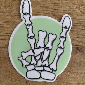 Peut inclure: Une main de squelette blanche faisant le geste de rock and roll, avec l'index et l'auriculaire tendus, sur un fond de cercle vert.
