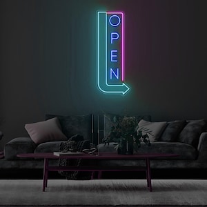 Könnte beinhalten: Ein Neon-Schild in Pfeilform, in den Farben Teal und Pink, zeigt das Wort "OPEN" in Blau. Das Schild ist an einer dunkelgrauen Wand über einem dunkelgrauen Sofa und einem weinroten Couchtisch angebracht.
