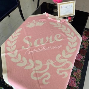 May include: A pink knitted baby blanket with a white crown, the name "Sare" and a snake design. The blanket has a white border and the text "GafullsBattaniye" is printed on the blanket.