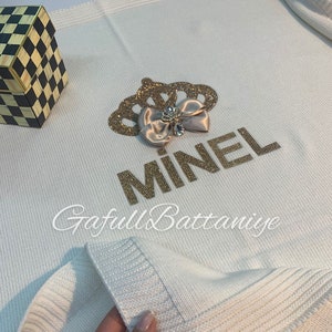 May include: White knitted baby blanket with a gold crown and the name "MINEL" embroidered in gold rhinestones. The blanket has a satin bow with a rhinestone in the center.