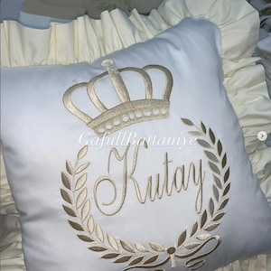 May include: White baby pillow with gold embroidery of a crown, laurel wreath, and the name "Kutay". The pillow has a ruffled edge and is part of a baby bedding set. The text "GafullBattaniye" is also visible.