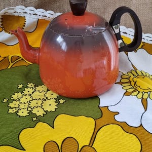 May include: A red and black enamel teapot with a black handle and a black knob on the lid. The teapot is sitting on a floral tablecloth with yellow, brown, and white flowers.