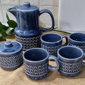 May include: A blue ceramic coffee set comprising a coffee pot, a lidded sugar bowl, and four mugs. Each piece features a repeating circle pattern around the base. The coffee pot has a lid and handle. The mugs have handles.