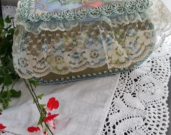 May include: A vintage-style trinket box with a green crocheted lid and a lace trim. The box features a colorful image of a cherub riding a carriage.