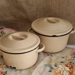 May include: Two cream-coloured enamel pots with lids. The pots have black trim and handles. The lids have a wooden handle. The pots are on a floral patterned tablecloth. The background is a burlap sack.