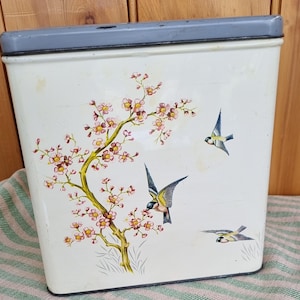Puede incluir: Un contenedor de almacenamiento de metal blanco vintage con tapa gris. El contenedor está decorado con una rama floral y tres pájaros en vuelo. Las flores son rosas y amarillas.