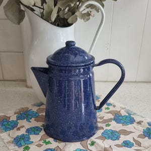 Puede incluir: Una cafetera de esmalte azul con un acabado moteado. La cafetera tiene una tapa y un asa. Está sentada sobre una tela con estampado floral.