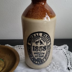 May include: A ceramic bottle with a cork stopper, featuring a cream-colored body and a brown glazed neck. The front label reads: MINERAL WATERS, POTTERS SINCE 1858, PINNACLE BRAND 1981, BENDIGO POTTERY AUSTRALIA, with an emu and kangaroo graphic.