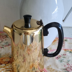 Puede incluir: Una cafetera dorada vintage con asa y tapa negras. La cafetera tiene una superficie brillante y reflectante y un pico para verter. Una jarra blanca está en el fondo. La cafetera está sobre una tela con estampado floral.