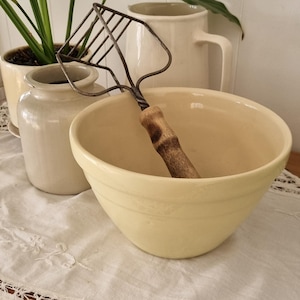 May include: A cream-coloured ceramic mixing bowl with a smooth finish, containing a metal potato masher with a dark, weathered wooden handle. The masher features a grid-like wire head and a twisted metal shaft connecting to the cylindrical handle.