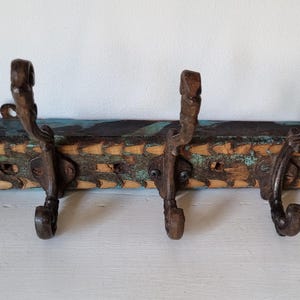 May include: A wall-mounted coat rack featuring a rectangular piece of weathered, reclaimed wood with visible teal and brown paint remnants. Three dark, ornate, cast-iron hooks are securely attached to the front, each featuring a curved design.