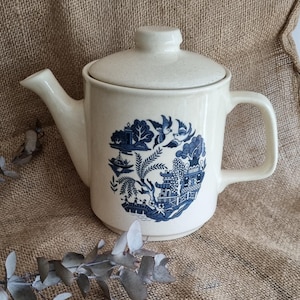 May include: Cream ceramic teapot with a lid and handle. The teapot has a blue and white Willow pattern design. The spout and handle are curved. The teapot is on a burlap surface.