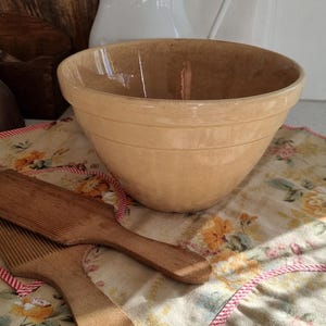 May include: A tan ceramic mixing bowl with a smooth interior and two horizontal ridges on the exterior, accompanied by two wooden butter paddles with ribbed surfaces, all resting on a floral patterned fabric with a red and white striped trim.
