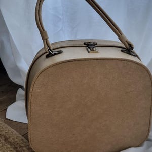 Pode incluir: Bolsa de mão vintage bege com alça curva e fecho de metal. A bolsa tem formato arredondado e superfície texturizada. Um acessório clássico.