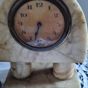 May include: A vintage marble clock with a round face and Roman numerals. The clock has a cream and brown color scheme, with a dark bronze-colored bezel. The clock is supported by two cylindrical legs and a rectangular base.