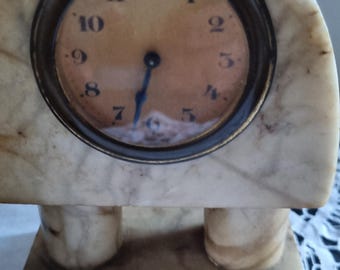 Könnte beinhalten: Eine antike Marmoruhr mit rundem Zifferblatt und römischen Ziffern. Die Uhr hat ein Creme- und Braunschema mit einer dunkelbronzenen Lünette. Die Uhr wird von zwei zylindrischen Beinen und einem rechteckigen Sockel getragen.