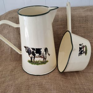 May include: A cream-coloured enamel pitcher and a matching ladle, both featuring a dark green rim and a graphic illustration of a black and white cow standing on a patch of green grass. The pitcher has a side handle and a pouring spout.