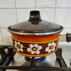May include: A brown and orange enamel saucepan with a black lid and a floral pattern. The saucepan has a white background with brown and orange flowers and green leaves. The saucepan is sitting on a black and silver stovetop.