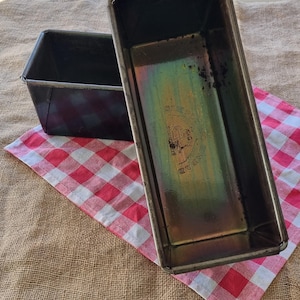 May include: Two rectangular metal loaf tins with a dark, weathered finish. The interior of the tins shows iridescent discolouration and a stamped logo that reads "SIMPLY BREAD" with a small graphic of a loaf of bread in the centre of the base.