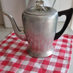 May include: A vintage aluminum coffee pot with a black handle and a clear glass lid. The coffee pot is sitting on a red and white checkered cloth. A white vase is in the background.