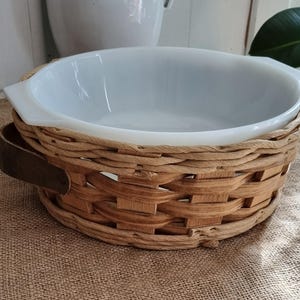 Puede incluir: Un plato para hornear de cerámica blanca con una base de cesta de mimbre tejido. El plato tiene una forma redondeada y un esmalte blanco liso. La cesta está hecha de mimbre marrón natural y tiene un patrón tejido.