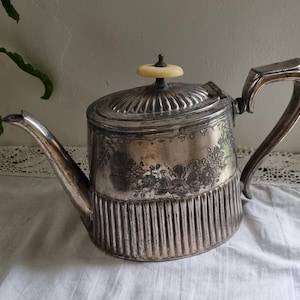 Peut inclure: Une théière en argent avec un design orné et un bouton crème sur le couvercle. La théière a un corps cannelé et un motif floral.