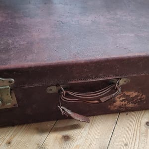 May include: A well-worn, rectangular suitcase in a dark burgundy hue. It features metal clasps, a leather handle, and a keyhole. The aged appearance suggests it's an antique, possibly made of leather, with signs of use.