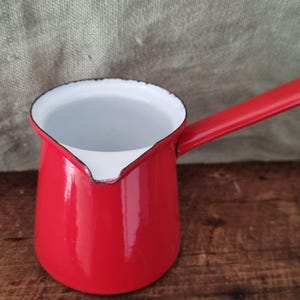 Puede incluir: Una pequeña olla cilíndrica de esmalte con un exterior rojo vibrante y un interior blanco liso. Cuenta con un mango largo único que se extiende desde el lateral y un pequeño pico vertedor integrado en el borde, con un ligero desgaste.