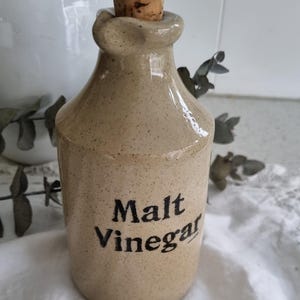 May include: A tan ceramic bottle with a cork stopper, labeled "Malt Vinegar" in black text. The bottle has a speckled finish and a rounded shape, suggesting a vintage or rustic style. The background includes a white pitcher and greenery.