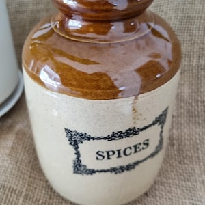 May include: A ceramic storage jar with a glossy brown glazed neck and a cream-coloured body. The jar features a black rectangular decorative border with the word SPICES printed in the centre. It has a wide, rounded opening and a cylindrical shape.